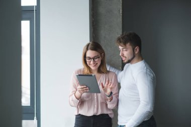 Pozitif genç Ofis çalışanları ofis duvar arka plan üzerinde duran ve tablet kullanma. Bir adam ve bir kız tablet vasıl seyir ve gülümseyen.