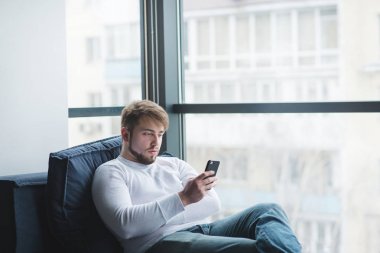 Pencereden kanepede oturan ve bir cep telefonu kullanarak bir yakışıklı sakallı bir adam. Sonra bir akıllı telefon kanepede oturan ofis çalışanı.