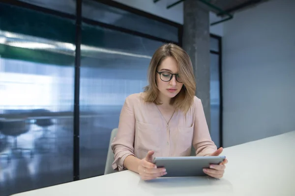 Όμορφη νεαρή γυναίκα με τα γυαλιά, κάθεται στο γραφείο του στο γραφείο και χρησιμοποιώντας ένα tablet. Κοιτάξτε το δισκίο. — Φωτογραφία Αρχείου