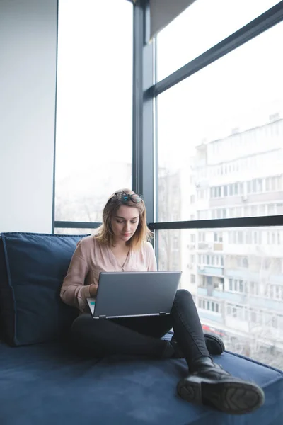Uma menina se senta em um sofá na janela em um escritório moderno e trabalha para um laptop. Trabalho em um escritório moderno . — Fotografia de Stock