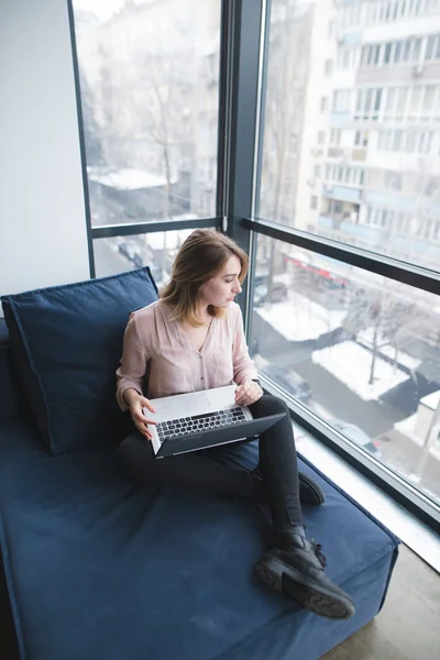 Empregada de escritório sentada em um sofá na janela e trabalhando em um laptop. Um olhar na janela. Trabalho em um escritório moderno . — Fotografia de Stock