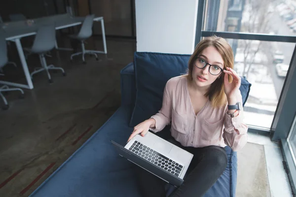 Retrato de uma menina bonita em óculos e um laptop. Um belo trabalhador de escritório posando na câmera enquanto se senta em um sofá com um laptop em seu colo. Um olhar para a câmera . — Fotografia de Stock