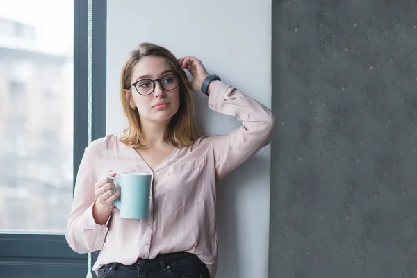 Beautiful girl posing near the wall in the office with a cup of coffee in his hands. Coffee break in the office. Weekdays office worker.