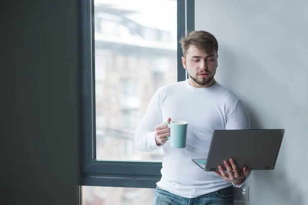 Um homem no escritório está junto à janela com um laptop e uma xícara de bebida quente em suas mãos. Pausa para café. Um homem trabalha de pé com uma xícara de café — Fotografia de Stock