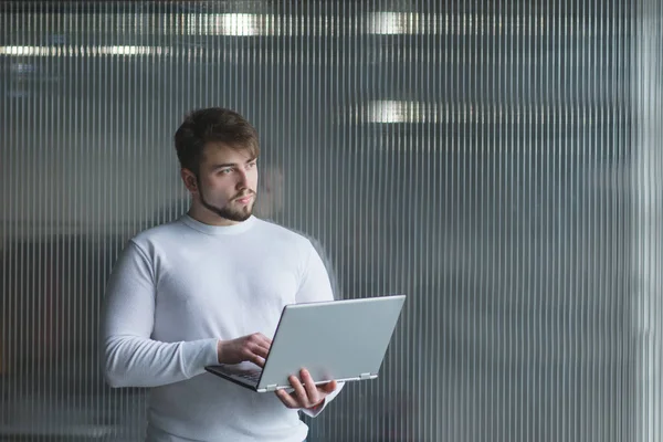 Красивый мужчина, стоящий с ноутбуком в руках на фоне офисной стены. Отвернись — стоковое фото