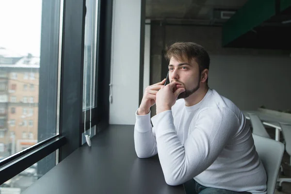 Un joven con barba se sienta en una oficina moderna en la mesa cerca de la ventana y habla por teléfono. El hombre habla por teléfono y mira por la ventana. — Foto de Stock