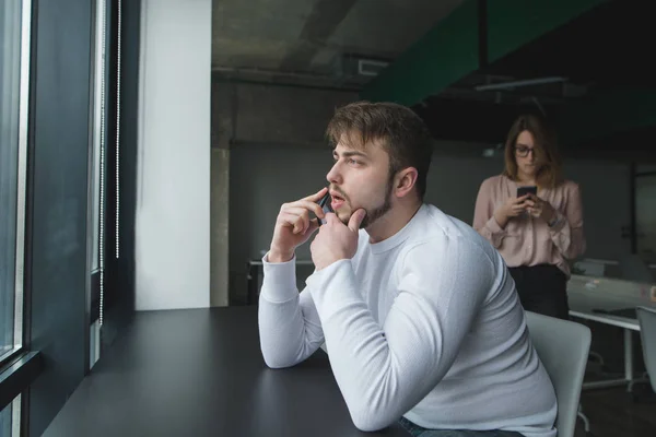 Un hombre serio está sentado en el escritorio de la oficina, hablando por un teléfono celular y mirando por la ventana. Trabajo en la oficina —  Fotos de Stock