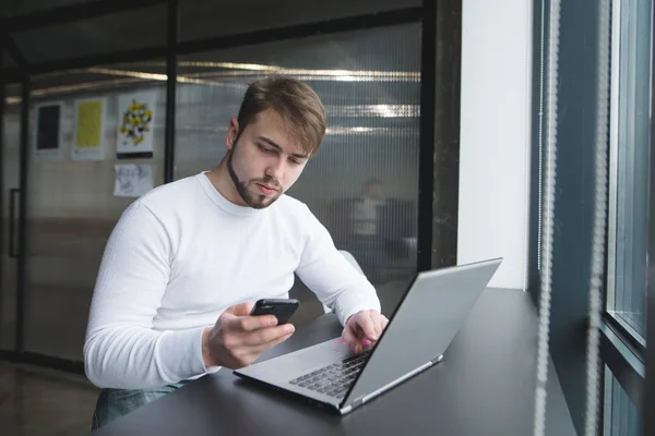 年轻男子坐在办公桌附近的笔记本电脑和使用手机。一个人在办公室里使用小玩意 — 图库照片