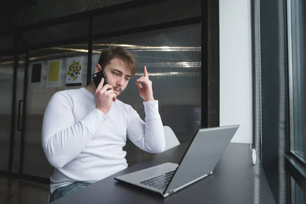 Un hombre serio está sentado en una oficina moderna cerca de la ventana en la mesa, y habla por teléfono freelancer trabaja en coworking . —  Fotos de Stock