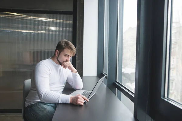 Freelancer trabaja en coworking. Un joven guapo trabaja en una computadora en una mesa cerca de la ventana. Trabajar en un portátil cerca de la ventana —  Fotos de Stock