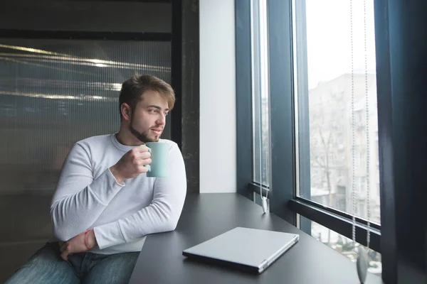 Un joven guapo sentado en una mesa con una taza de café cerca de un portátil cerrado durante un descanso en el trabajo. Un hombre en la oficina con una taza de bebida en las manos mira por la ventana . —  Fotos de Stock