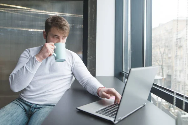 Joven trabajando en una laptop en coworking y bebiendo café. Un hombre bebe café mientras trabaja en la oficina cerca de la ventana —  Fotos de Stock