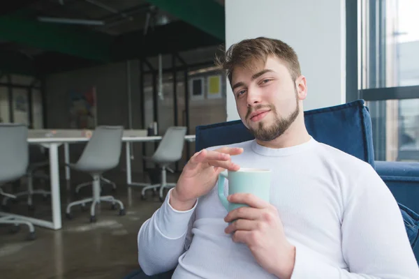 Un hombre guapo se sienta en la oficina con una taza de café en sus manos y sonríe. En el descanso, el oficinista bebe una bebida caliente de la taza. —  Fotos de Stock