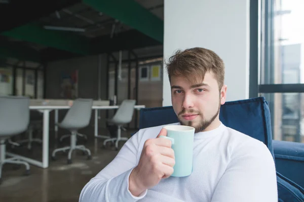 Retrato de un joven con una taza de bebida en las manos al fondo de la oficina. Un hombre con una taza de café descansando en la oficina en el sofá . — Foto de Stock