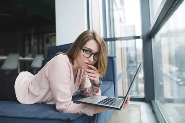 Potret seorang gadis cantik di kantor di sofa dengan laptop di tangannya. Seorang pekerja kantor bekerja pada laptop di sofa dan melihat ke kamera — Stok Foto