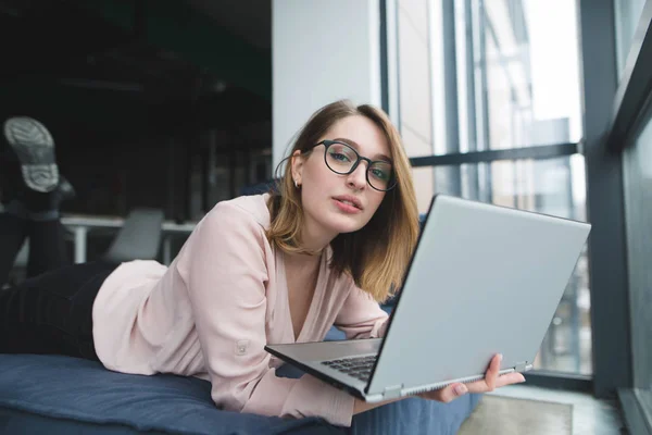 Uma menina bonita nos óculos fica na janela do sofá com um laptop nas mãos e olha para a câmera. O freelancer trabalha em um coworking deitado no sofá — Fotografia de Stock