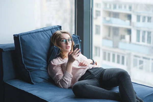 Güzel bir kız ofis pencereden kanepede yatıyor ve bir akıllı telefon kullanır. Kız kanepede oturan ve kameraya bakıyor — Stok fotoğraf