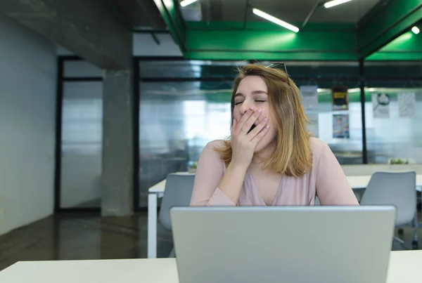The office worker sits at the office in the office behind a laptop and yawns. A tired girl works for a computer in a modern office