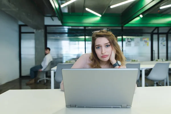 Uma menina triste com uma xícara de café nas mãos usa um laptop na mesa no escritório. Uma menina cansada trabalha para um laptop e bebe uma xícara de café — Fotografia de Stock