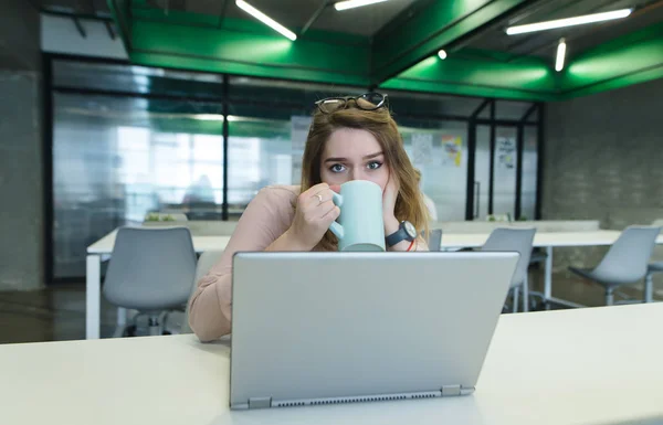 Gyönyörű lány ül az irodában a pultnál, és kávét inni a poharat, egy számítógépen dolgozva. Irodai munka fogalma. — Stock Fotó
