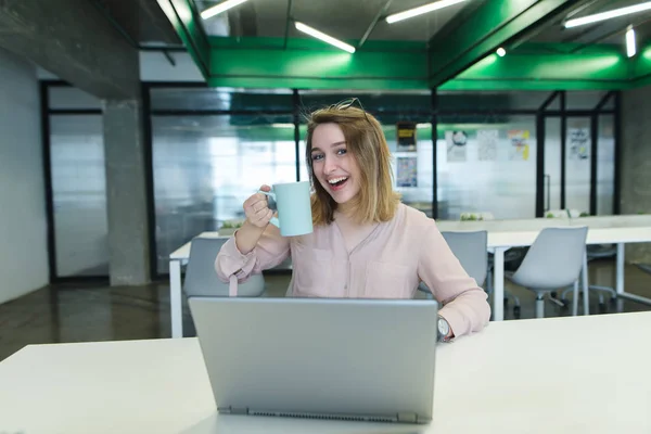 Menina sorrindo com uma xícara de café em seus braços sentado na mesa no escritório e usando um laptop. Pausa para café durante o trabalho . — Fotografia de Stock