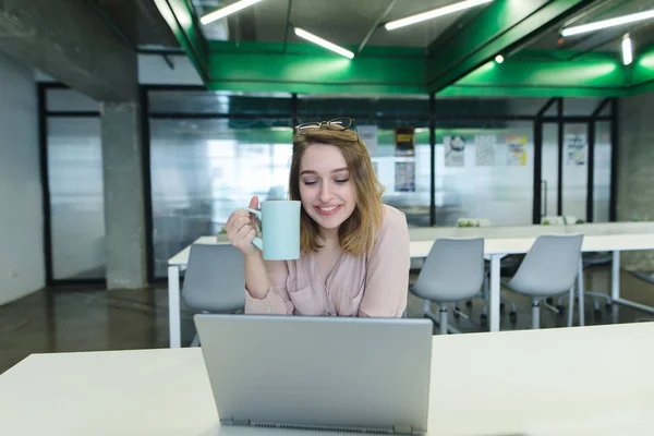 Menina sorridente, um funcionário de escritório com uma xícara de café em seus braços sentado à mesa e trabalhando em um laptop. Trabalho com laptop e café . — Fotografia de Stock