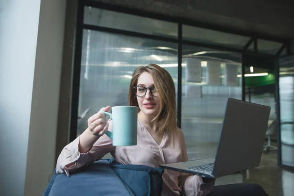 그녀의 손에서 음료 한잔과 함께 아름 다운 소녀는 노트북에 작업 하는 동안 카메라에 보인다. 커피 한 잔 함께 소파에 작동 하는 여자 프리랜서 — 스톡 사진