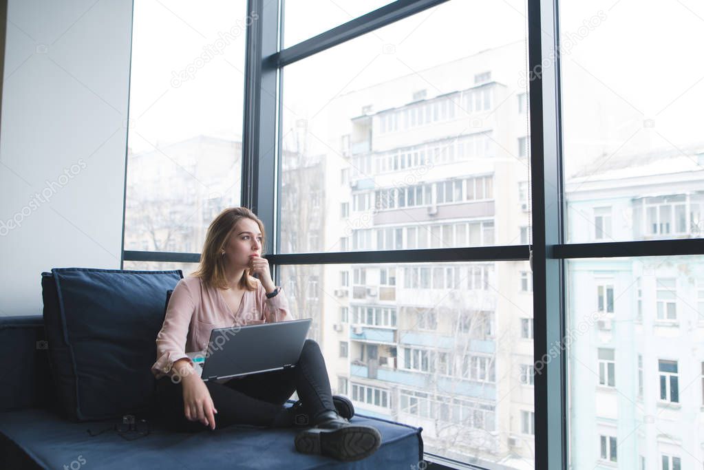 Beautiful girl sitting on a sofa with a laptop on her lap and looking at the panoramic window. Work in a modern office on a sofa.