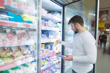 A man chooses frozen foods from shelves in a refrigerator in a supermarket. A man buys products in the store. Shopping in a supermarket concept. clipart