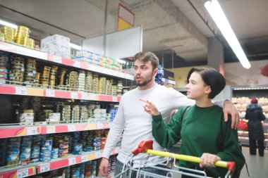 Genç bir çift bir süpermarket alışveriş sepeti ile size yürür, raf görünüyor ve mal seçer. Bir süpermarkette alışveriş aile