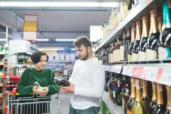 A young beautiful couple buys alcohol in the store. A couple chooses wine at a supermarket. — Stock Photo, Image