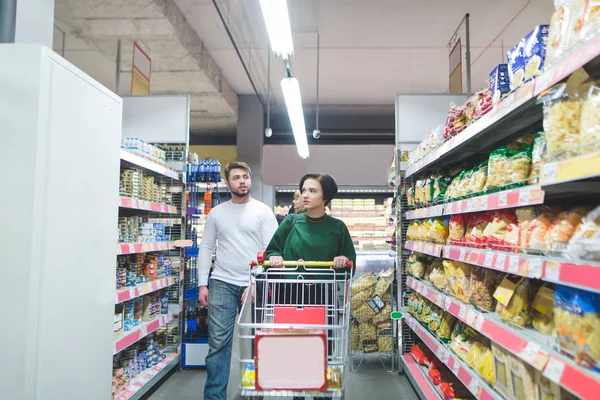 Giovane coppia a piedi attraverso il supermercato con un carrello e seleziona i prodotti per lo shopping. Shopping in famiglia in un supermercato — Foto Stock