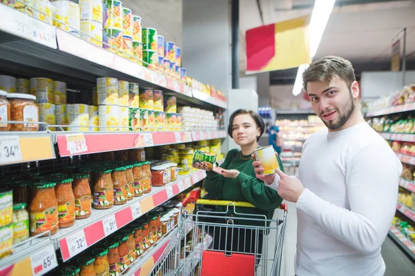 Fiatal gyönyörű pár pózol egy szupermarket a termékek a kezükben. Egy fiatal férfi és egy lány vesz egy szupermarketben konzerv zöldségek — Stock Fotó