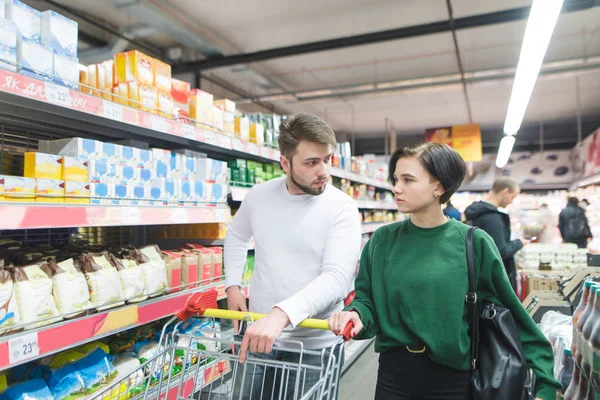Una hermosa pareja joven caminando en un supermercado con un carro y hablando. Selección familiar de productos de supermercado —  Fotos de Stock