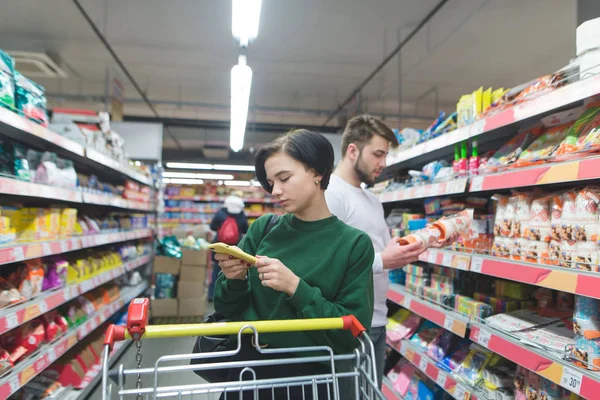 Des jeunes au supermarché. Une fille regarde son smartphone pendant ses achats, et son mari lit l'étiquette du produit. Shopping dans un concept de supermarché — Photo