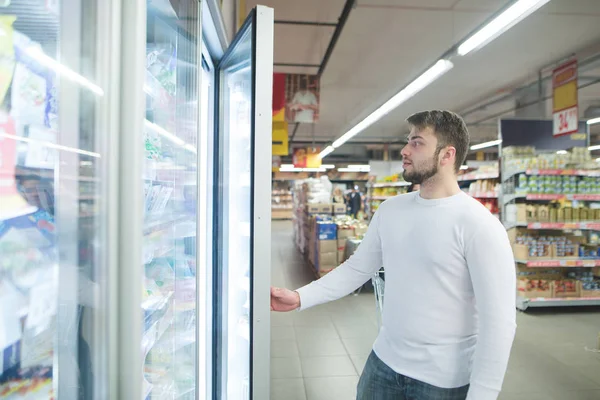 Um belo barbudo abre um frigorífico num supermercado. Compras no supermercado — Fotografia de Stock