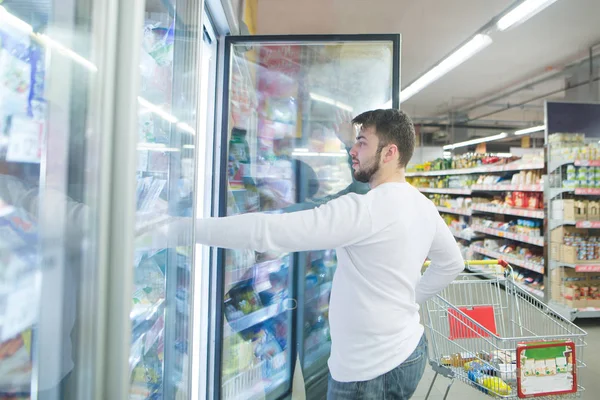 Un joven con barba toma alimentos congelados de un refrigerador en un supermercado. Compras en la tienda — Foto de Stock