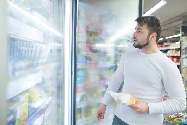 Retrato de un hombre cerca de un refrigerador en un supermercado con alimentos congelados en sus manos. Compra de productos en un supermercado . — Foto de Stock