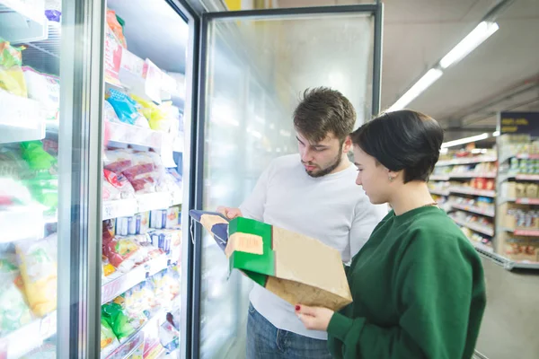 A young couple of buyers are looking at frozen foods near the refrigerator at a supermarket. Family shopping at a supermarket. A look at the label of the product. — Stock Photo, Image