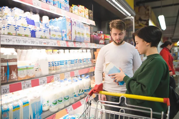 Piękna Młoda para wybiera produkty mleczne w supermarkecie. Dziewczyna czyta etyki mleka w sklepie. Rodzinne zakupy w supermarkecie — Zdjęcie stockowe