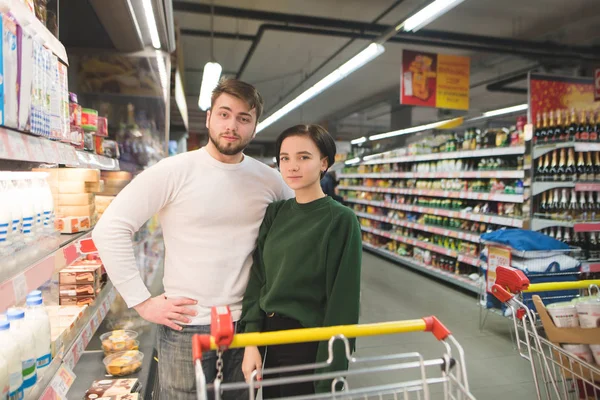 Joven hermosa pareja con carritos de compras de pie en el supermercado y mirando a la cámara. Compras familiares en un supermercado . — Foto de Stock