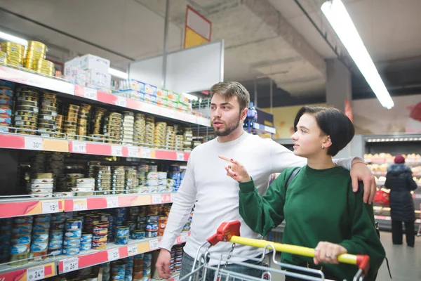Молодая пара проходит через супермаркет с тележкой, смотрит на полку и выбирает товар. Семейные покупки в супермаркете — стоковое фото
