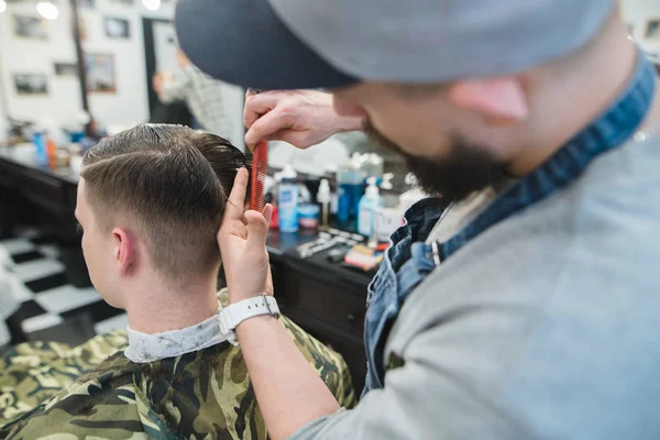 Barbeiro corta a tesoura e pente de um jovem nos barbercos. Cortes de cabelo masculinos no salão de beleza — Fotografia de Stock