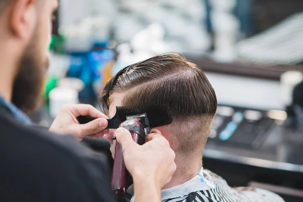Barbeiro com barba corta um jovem com um cortador de cabelo no salão de beleza masculino. Crie um penteado elegante na barbearia — Fotografia de Stock