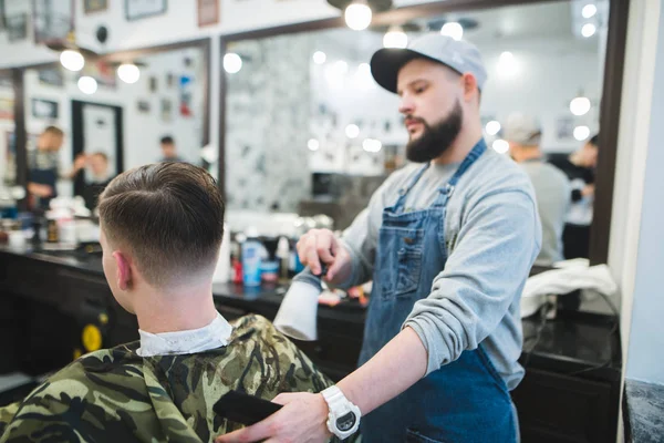Стильный парикмахер с бородой заканчивает мужскую прическу для клиента. Мужская стрижка в парикмахерской. Молодой красивый парикмахер стрижет привлекательного мужчину в парикмахерской — стоковое фото