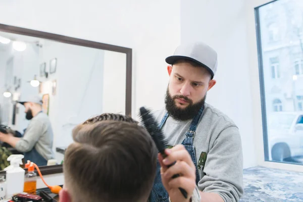 Barbier avec une barbe crée une coiffure élégante pour un jeune homme dans un salon de coiffure. Un coiffeur sérieux coupe un homme dans un salon de beauté pour hommes — Photo