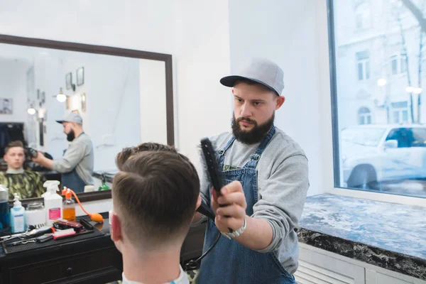 Cortes de cabelo masculinos na barbearia. Um cabeleireiro elegante com barba cria um penteado para um cliente no salão de beleza masculino. — Fotografia de Stock