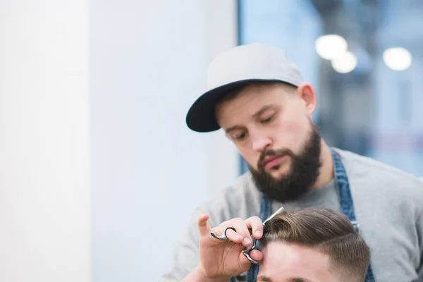 Ritratto di barbiere al lavoro. Un uomo con la barba taglia le forbici di un cliente nel salone di bellezza maschile. Crea un'acconciatura maschile alla moda — Foto Stock