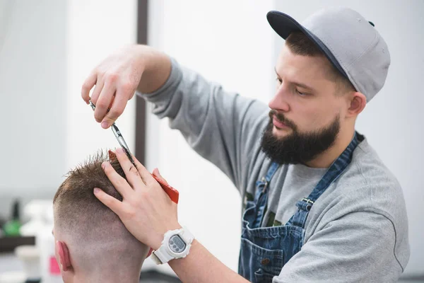 Portrait of a young barber who shaves a young man's hair scissors. A stylish hairdresser with a beard creates a hairstyle for a young man. — Stock Photo, Image