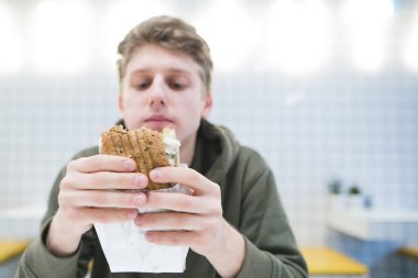 Aç genç adam elinde bir hamburger görünüyor. Öğrenci hafif bir restoranda şık iç mekanlara ile fast food yiyor